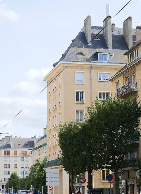 Immeuble d'habitation en pierre claire à Caen, avec toit d'ardoise, lucarnes et balcons, derrière une rangée d'arbres
